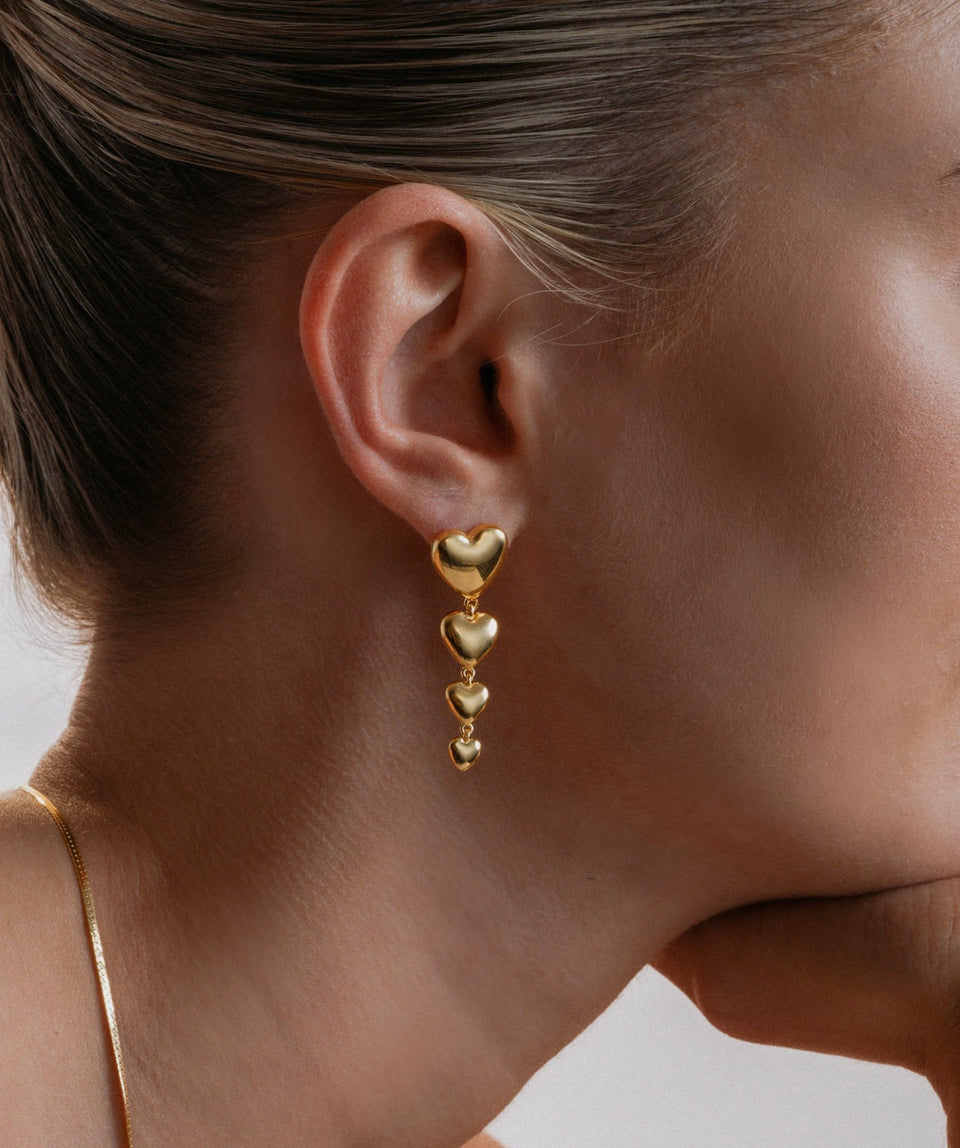 A close-up of a model wearing the Darling Earrings in gold, which feature a cascade of four bold hearts, paired with a delicate gold snake chain necklace.