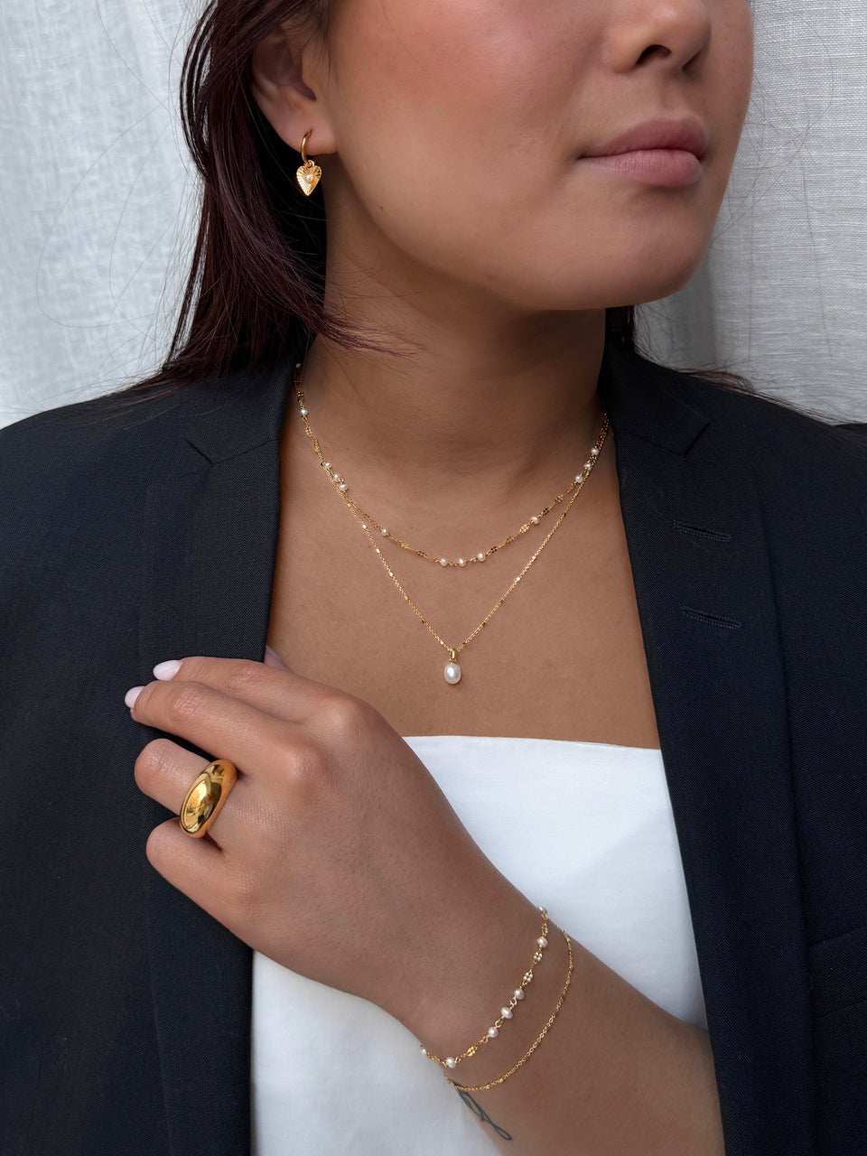 A close-up of a model wearing a complete gold Syster P jewelry set, featuring the layered Aurelia and Treasure pearl necklaces, Aurelia heart earrings, layered Aurelia and Sheer bracelets, and a Bolded Big Ring.