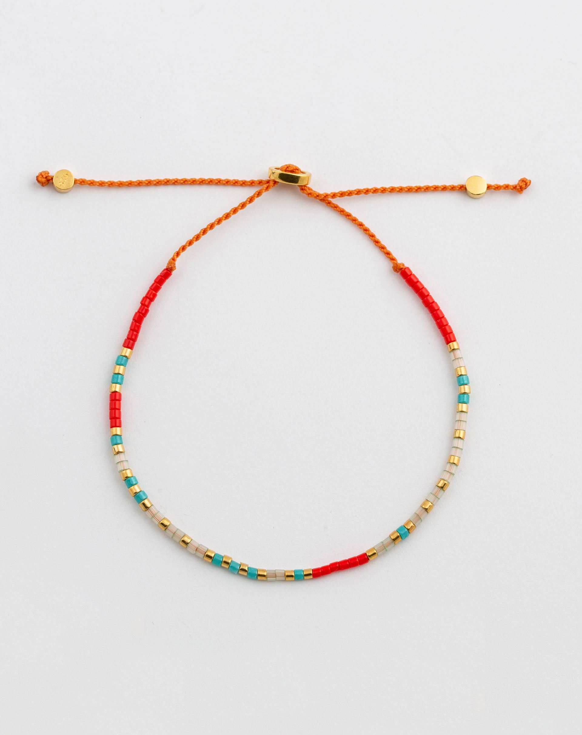 A product shot of the Code Bracelet Gold 'I Love You', featuring an adjustable orange cord and a pattern of red, turquoise, clear, and gold Japanese glass beads that spell out the message in Morse code.