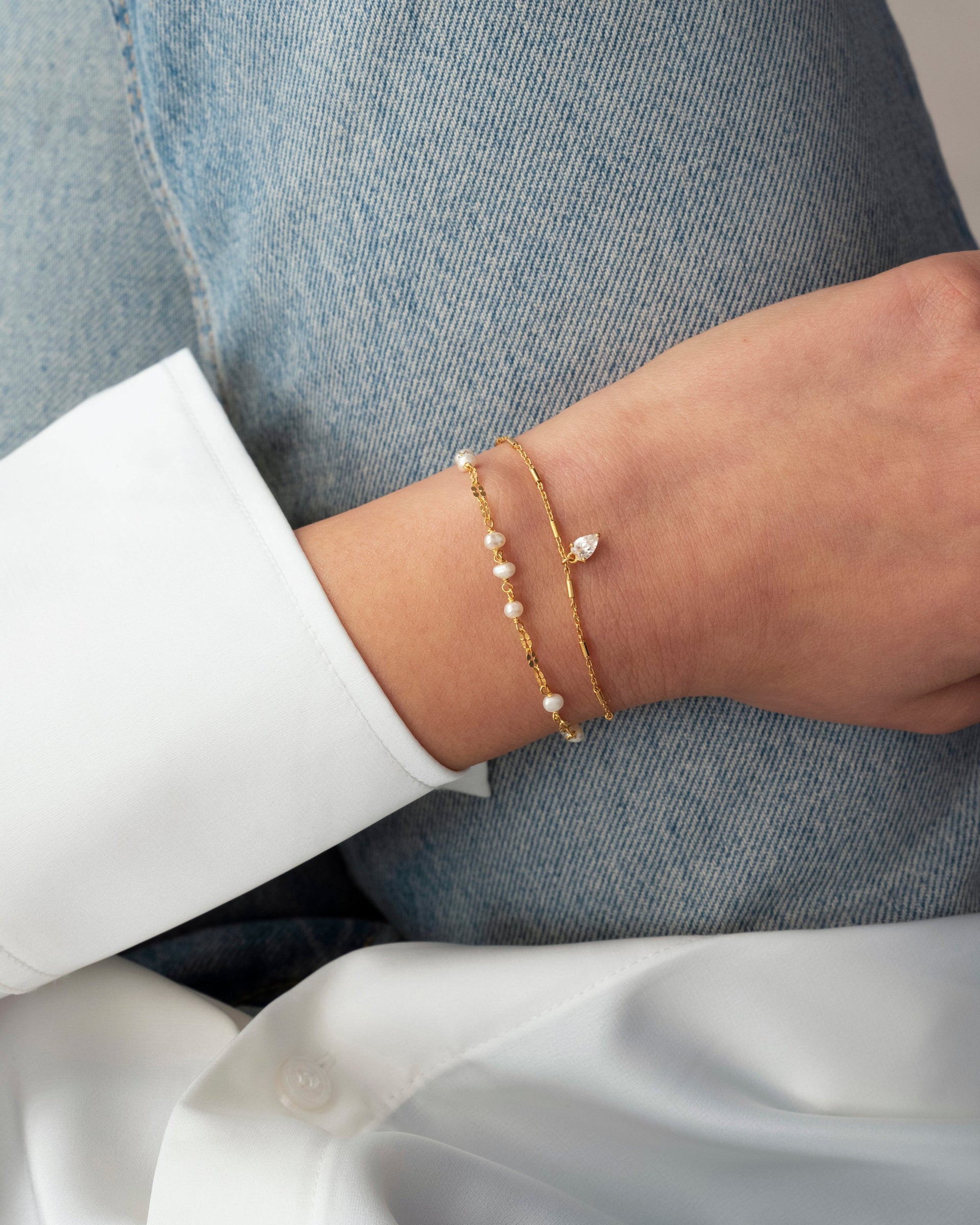 A close-up of a model's wrist wearing the Aurelia Bracelet in gold, which features small freshwater pearls, stacked with the Katie Bracelet, a fine gold chain with a teardrop-shaped crystal.