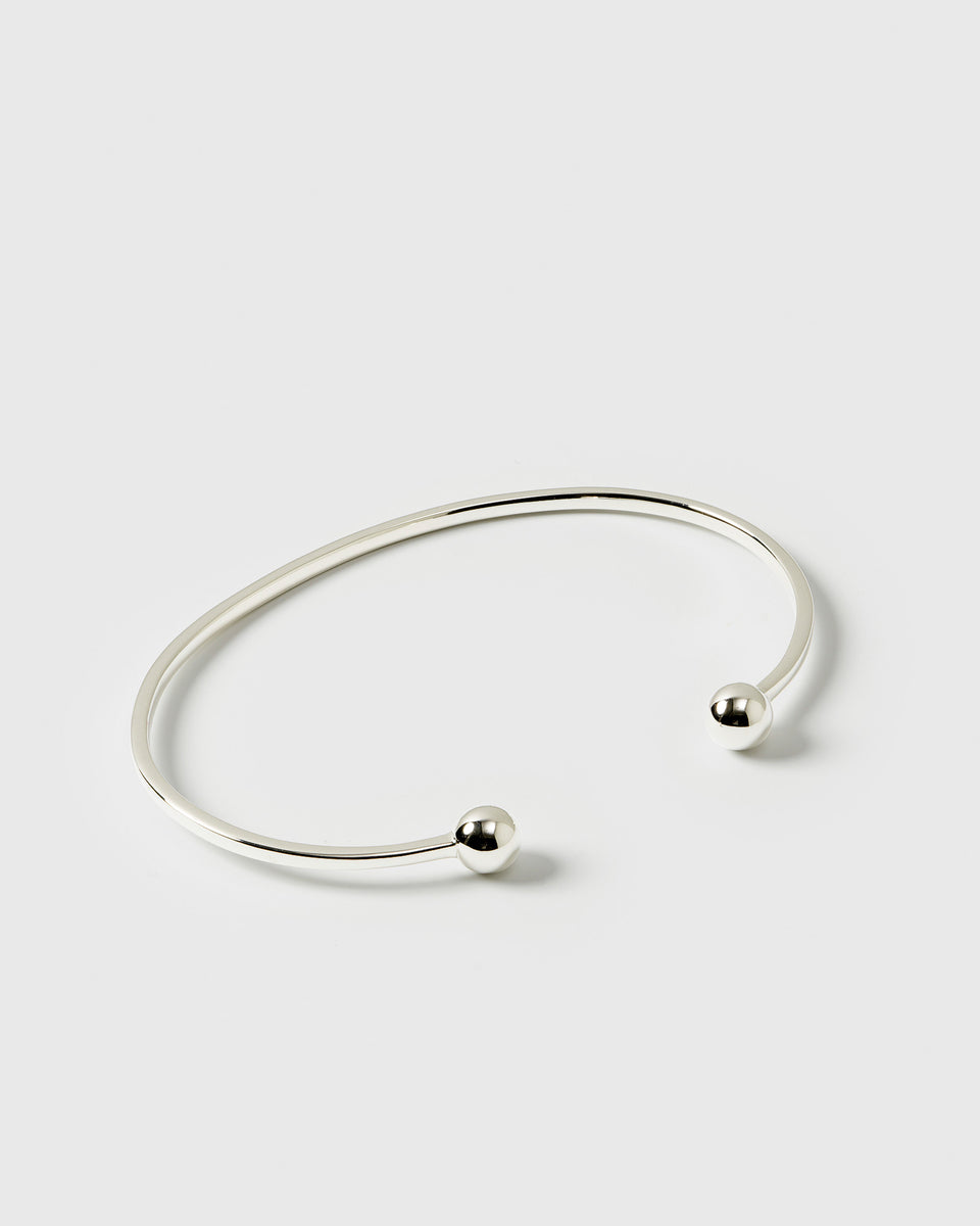 A product shot of the Strict Ball Bangle in silver, a thin, open cuff bracelet with a polished finish and small, rounded balls at each end.