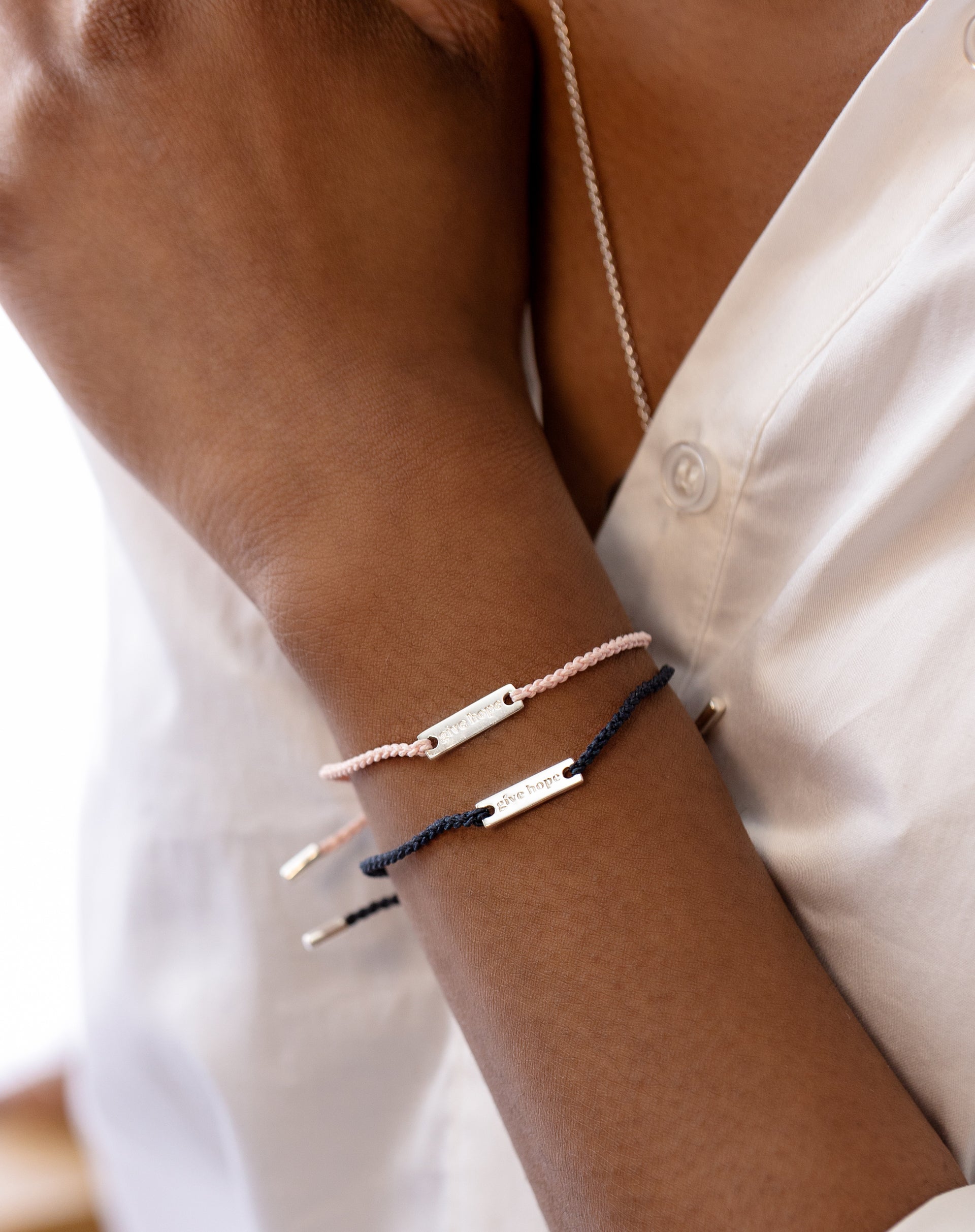 A model wears the Syster P Give Hope bracelet in navy blue stacked with a light pink version. Both thread bracelets feature a sterling silver plaque engraved with the words 'give hope'. A silver chain necklace is also visible.