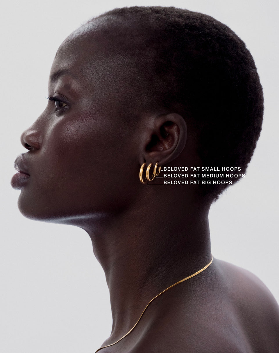 A model in profile wearing three sizes of the Beloved Fat Hoops in gold‚ small, medium, and big‚ stacked in her ear, paired with a delicate gold chain necklace.