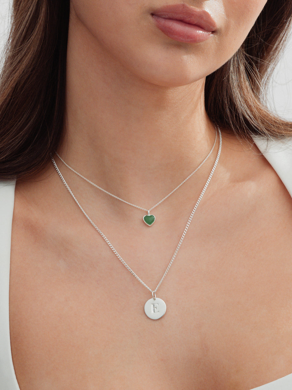 Close-up of a model wearing two layered silver necklaces: the La La Love necklace with a small green jade heart pendant, layered with a Beloved Short Curb Chain necklace with a round letter E pendant.