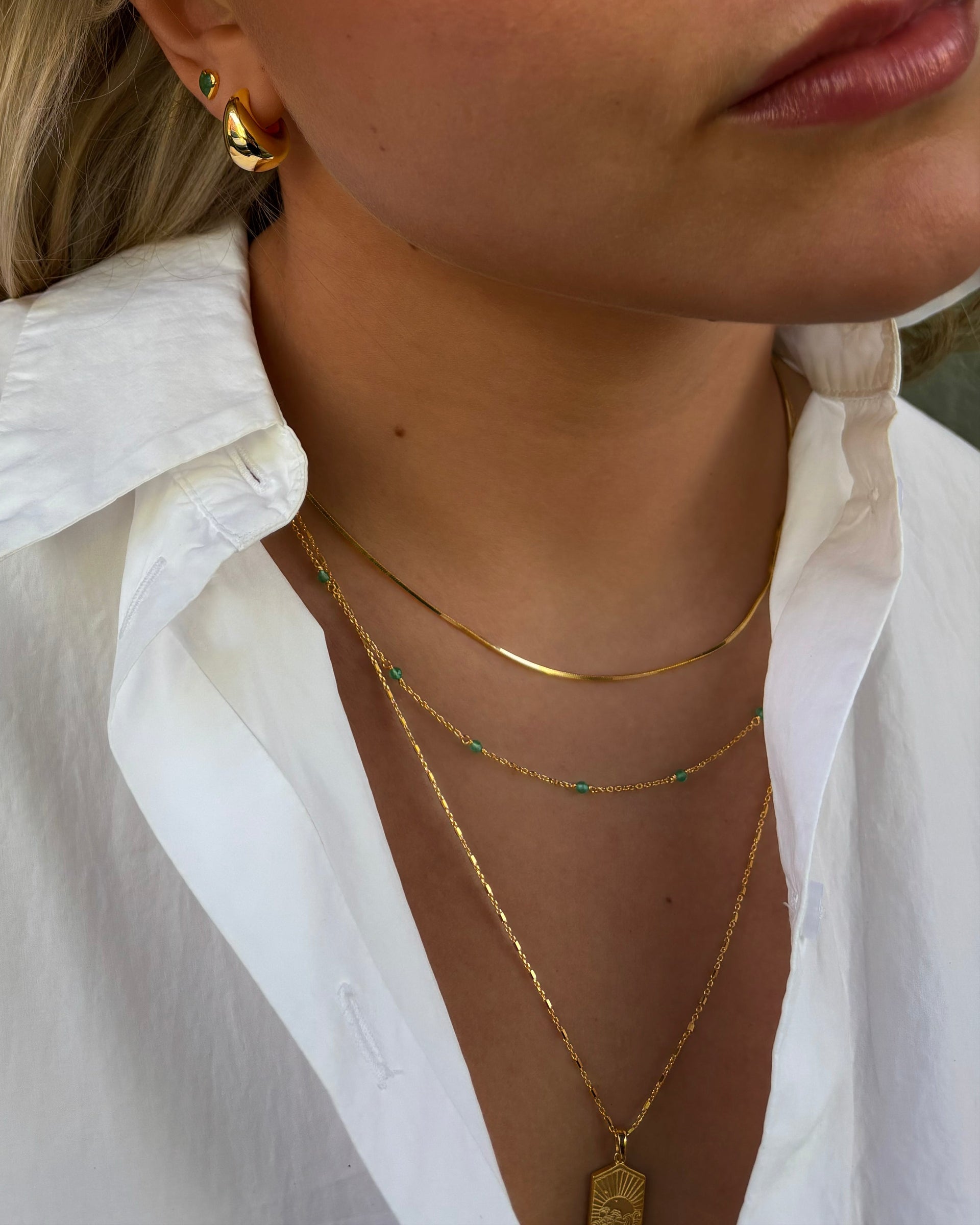 Close-up of a model wearing the Santa Monica Necklace Gold, a delicate gold chain with green onyx beads, layered with a gold chain and a gold pendant necklace, complemented by bold gold earrings and a green heart-shaped stud.
