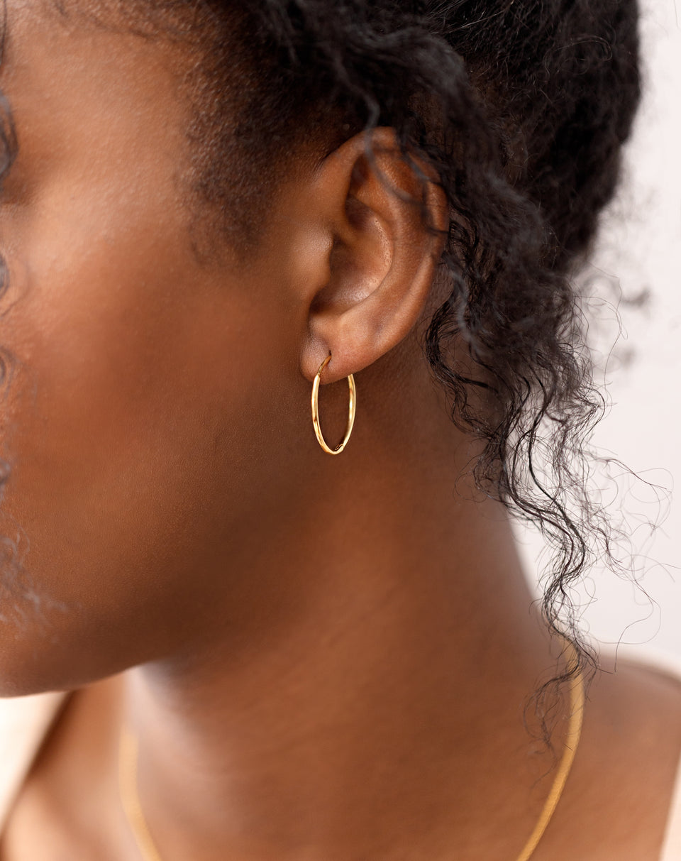 A close-up shot of a model wearing the classic Beloved Medium Hoops in gold, with a delicate gold chain necklace also visible.