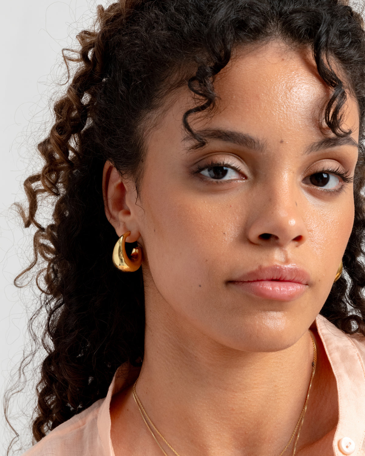 A close-up shot of a model wearing the Bolded Earrings in gold, highlighting their unique, chunky shape. She is also wearing a thin gold chain necklace.