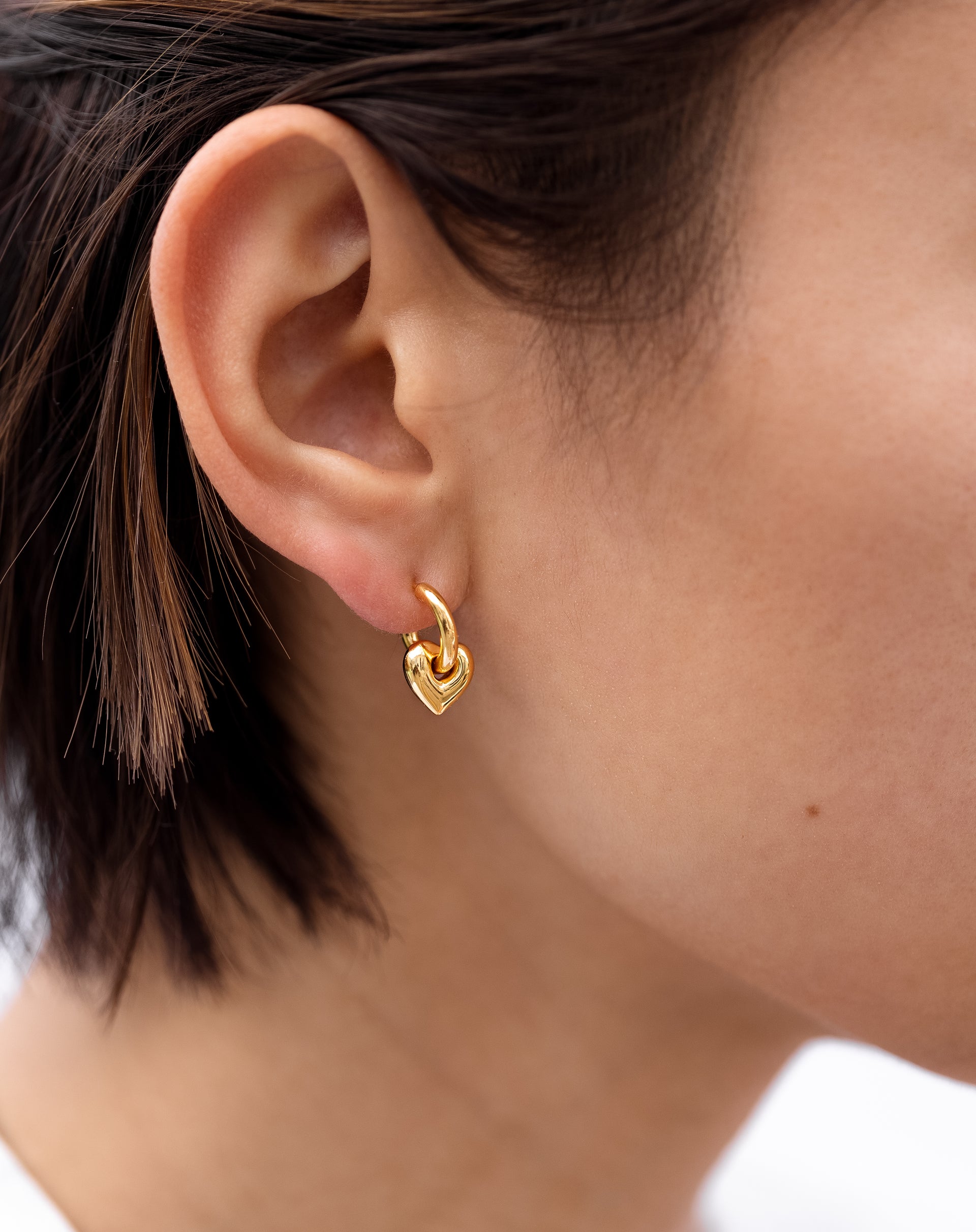 A close-up shot of a model wearing the La La Love Fat Heart Earrings in gold, featuring a small hoop with a chunky heart charm.