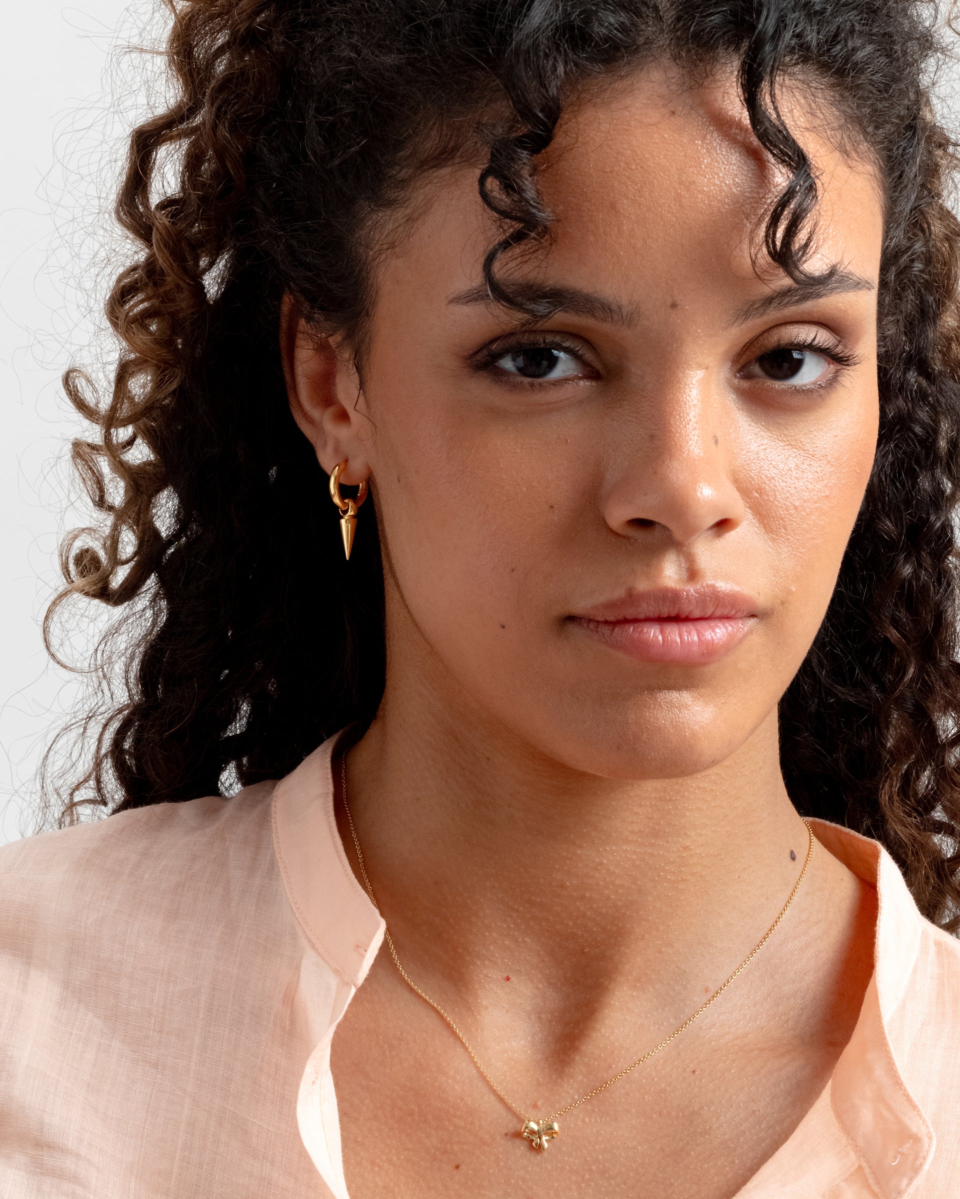 A close-up shot of a model wearing the Juno Single Hoop Gold, featuring a dangling spike charm, and a delicate gold necklace with a bow pendant.