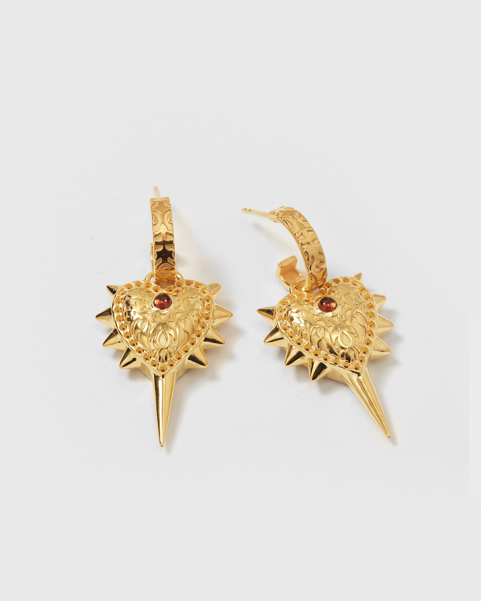 A product shot of the Drama Queen Heart Spike Earrings in gold, featuring patterned hoops with dangling, engraved heart pendants decorated with spikes and a small red stone.