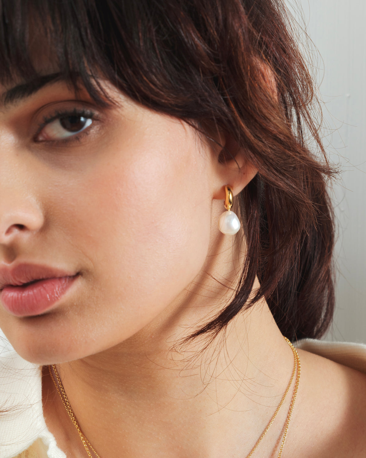 A close-up shot of a model wearing the Paxton Earrings in gold, featuring a dangling baroque freshwater pearl.