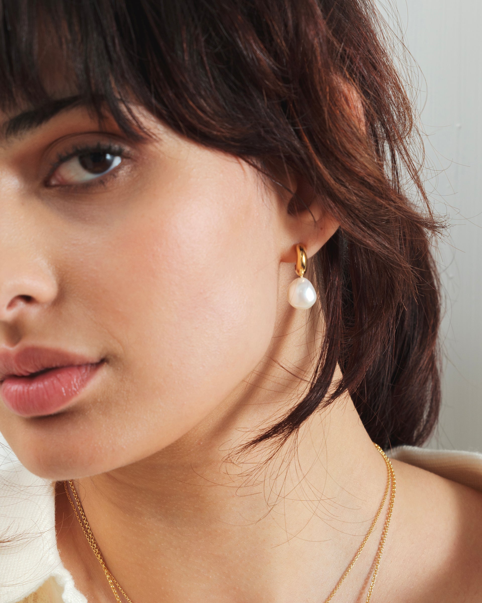A close-up shot of a model wearing the Paxton Earrings in gold, featuring a dangling baroque freshwater pearl.