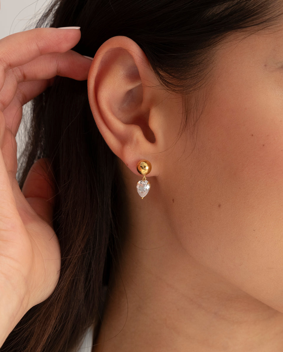 A model wears the Katie Earrings in gold, shown in a close-up to highlight the drop-shaped cubic zirconia that dangles from a polished gold stud.