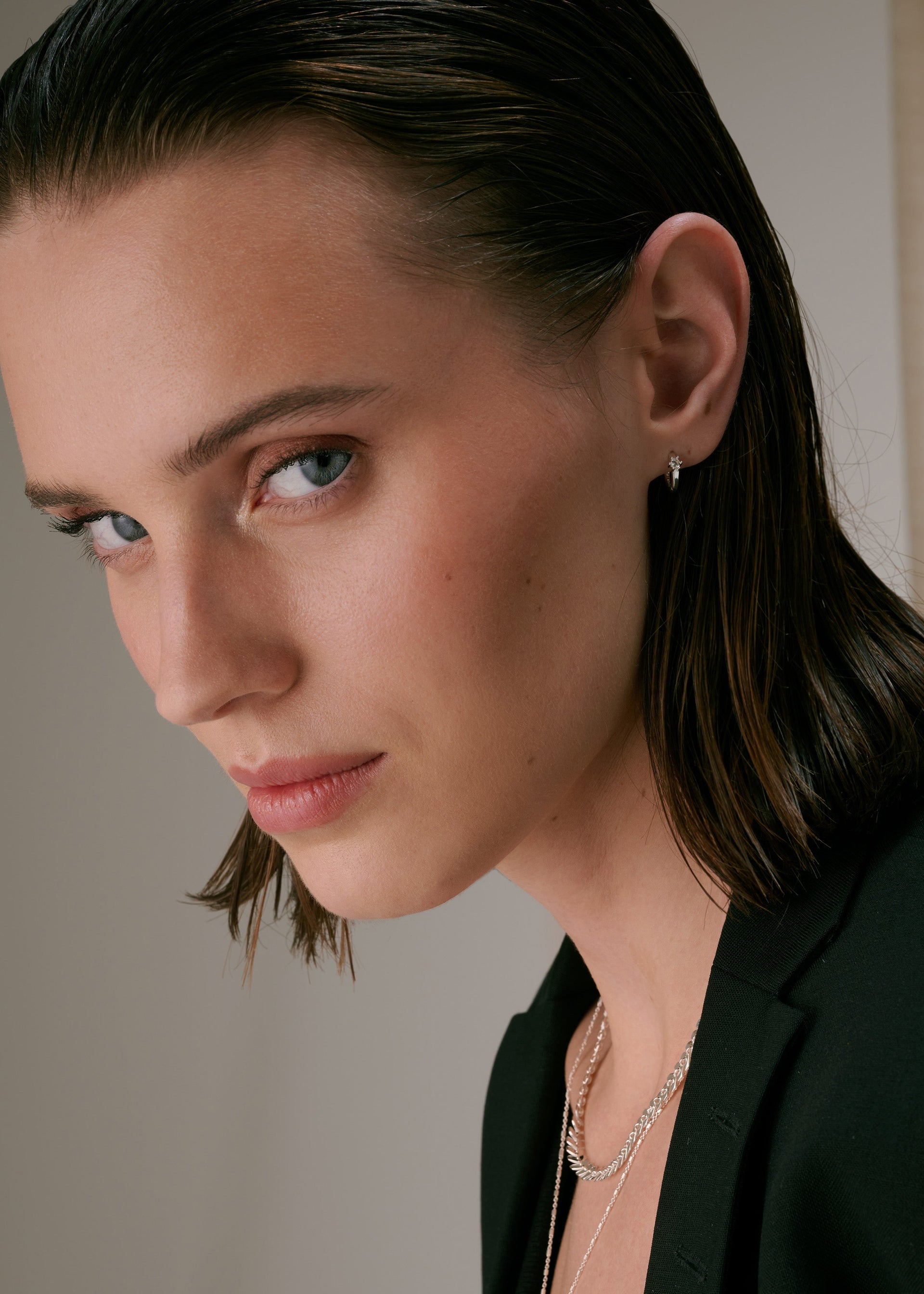 A close-up of a small silver hoop earring with a crystal, worn with several layered silver necklaces.