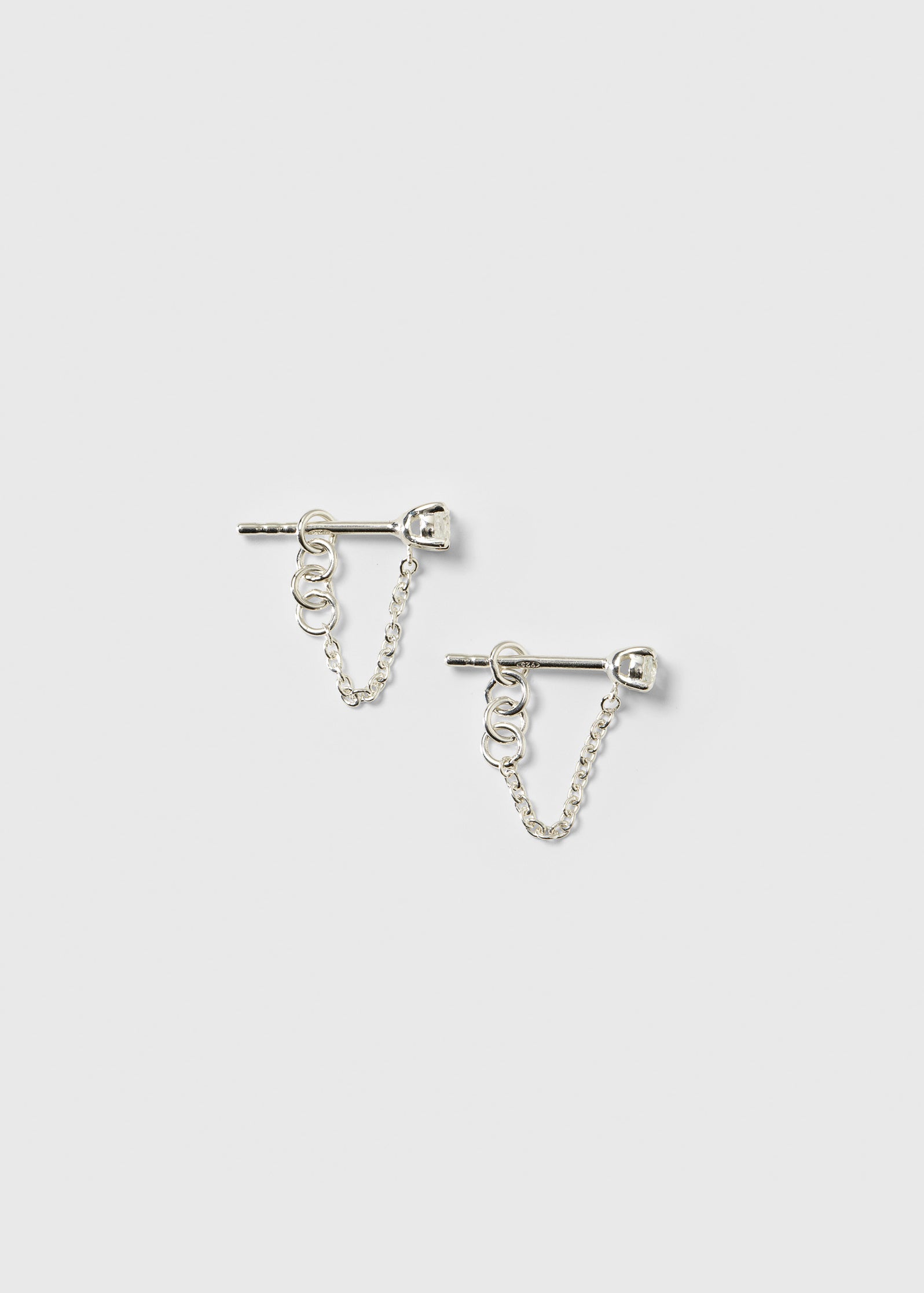 A product shot of the Minimalistica Solo Nova Earrings in silver, featuring a stud design with a white topaz and a delicate dangling chain.