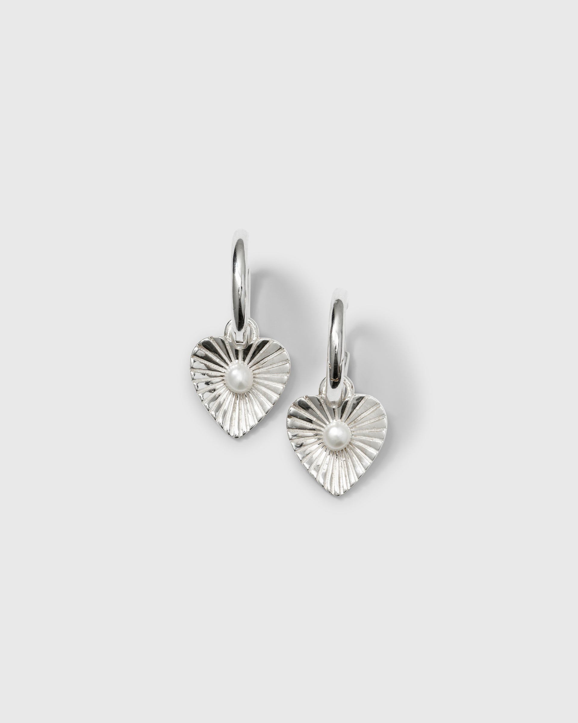 A product shot of the Aurelia Earrings in silver, featuring hoop earrings with a textured, heart-shaped pendant and a small freshwater pearl at its center.