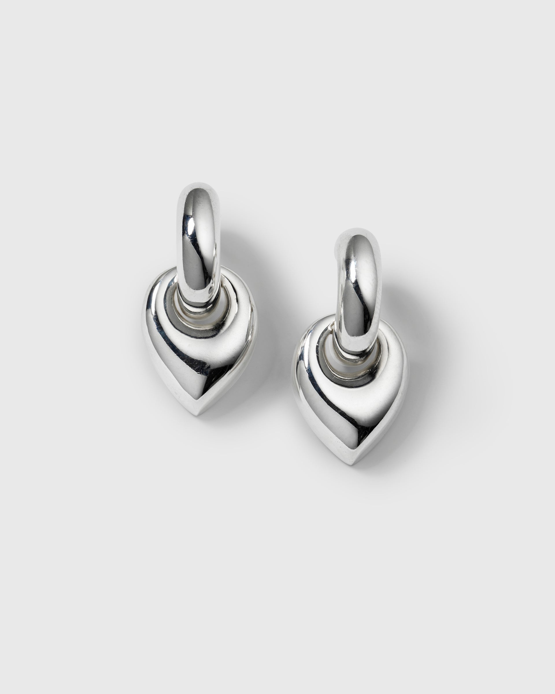 A product shot of the Melinda Earrings in silver, showcasing the pair's chunky hoop design with a dangling, leaf-shaped pendant.