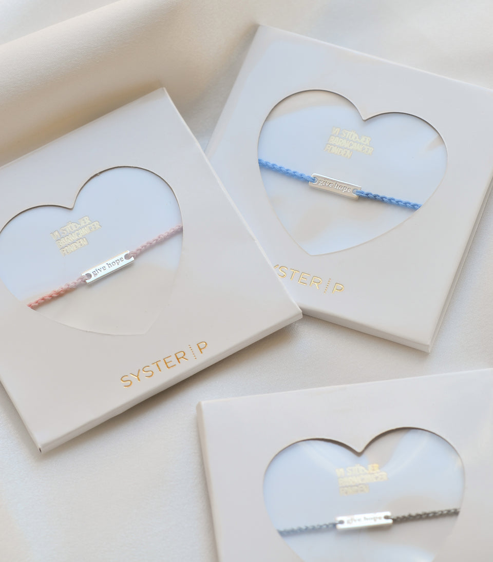 A product shot of the Syster P Give Hope bracelets in pink, blue, and silver metallic thread, each presented in its white box with a heart-shaped window to showcase the silver "give hope" plaque.