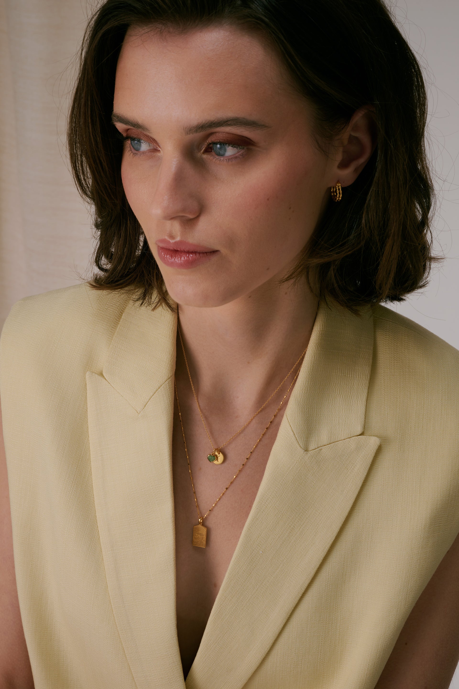 Layered gold necklace set with Mini Letter E pendant and green heart charm on top chain, rectangular pendant on bottom chain, paired with small textured gold hoop earrings.