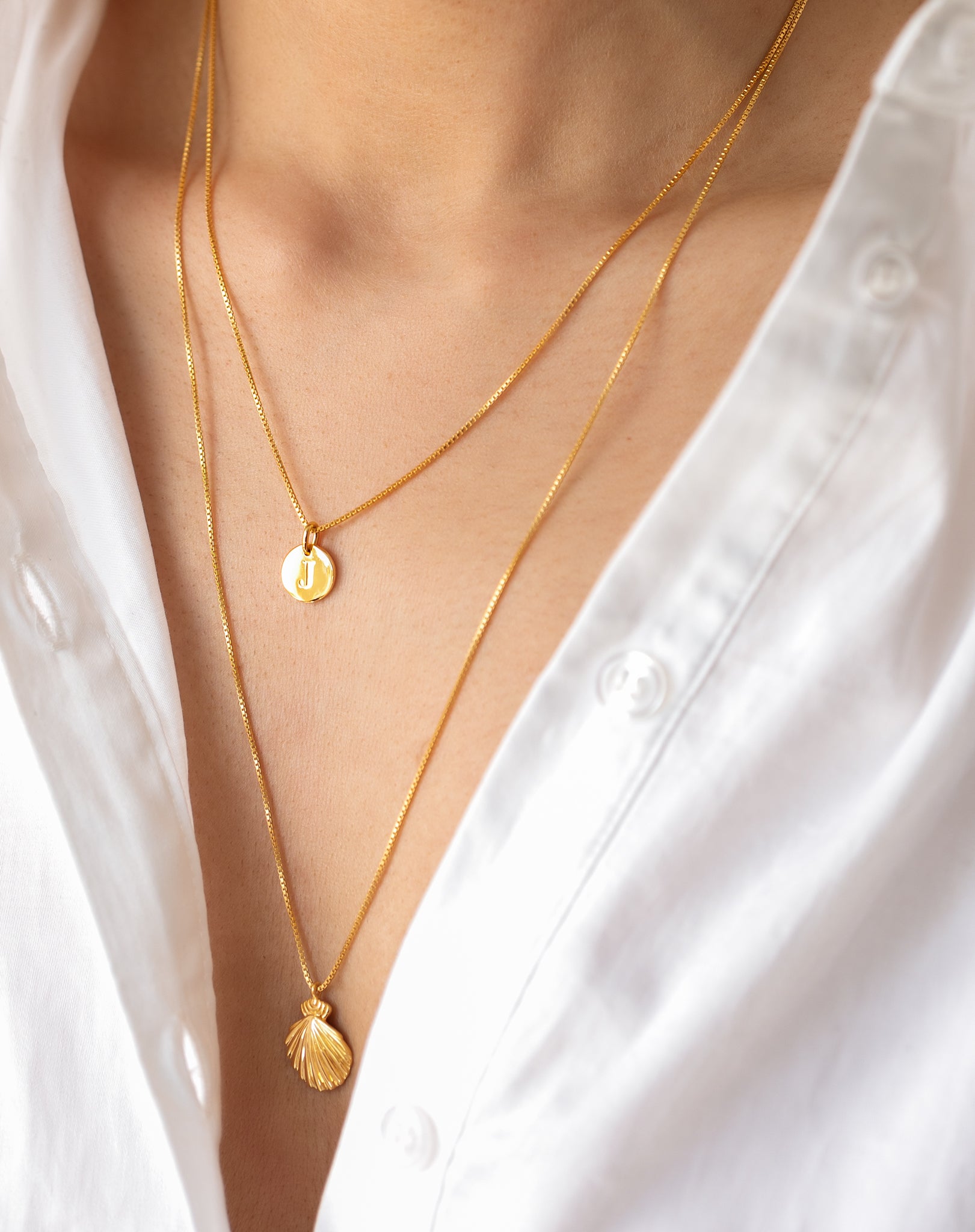 A model wearing the Beaches Shell Necklace in gold, featuring a shell-shaped pendant, layered with a shorter gold necklace with a round initial pendant.