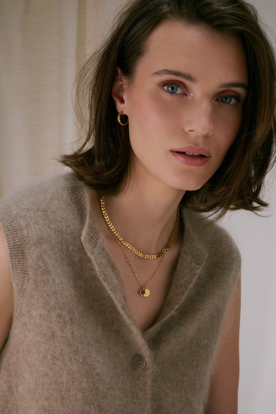 A gold necklace with a leaf-like pattern, layered with a delicate gold chain with charms, paired with a twisted gold hoop earring.