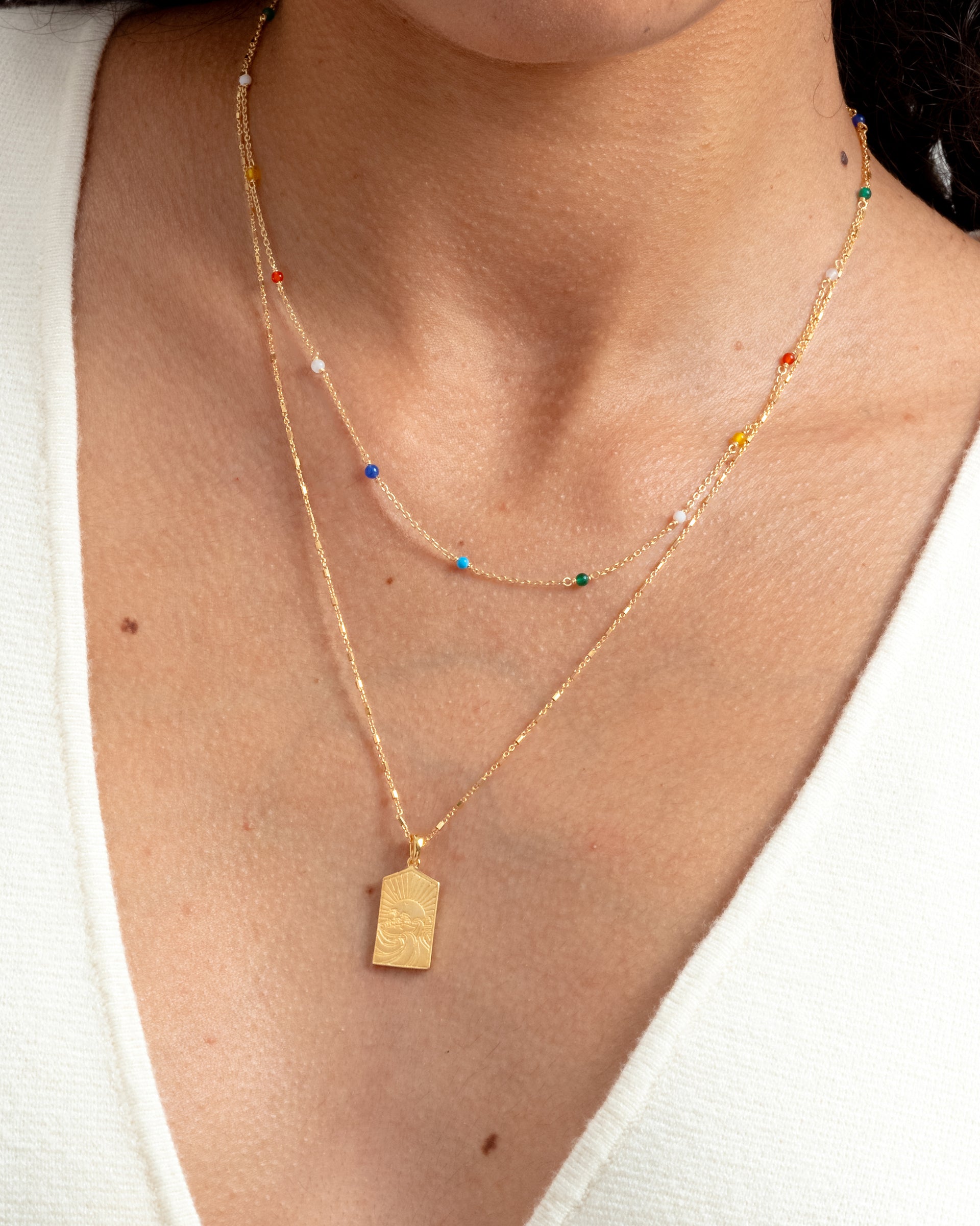 A model wears the Santa Monica Necklace in gold, a fine chain with colorful stones, styled in a layered look with another gold necklace featuring a rectangular sun pendant.
