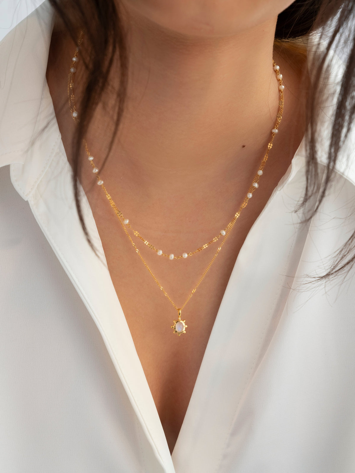 A close-up of a model wearing the gold Aurelia Necklace, featuring a delicate chain with small freshwater pearls, layered over a second gold necklace with a sun-shaped pendant.