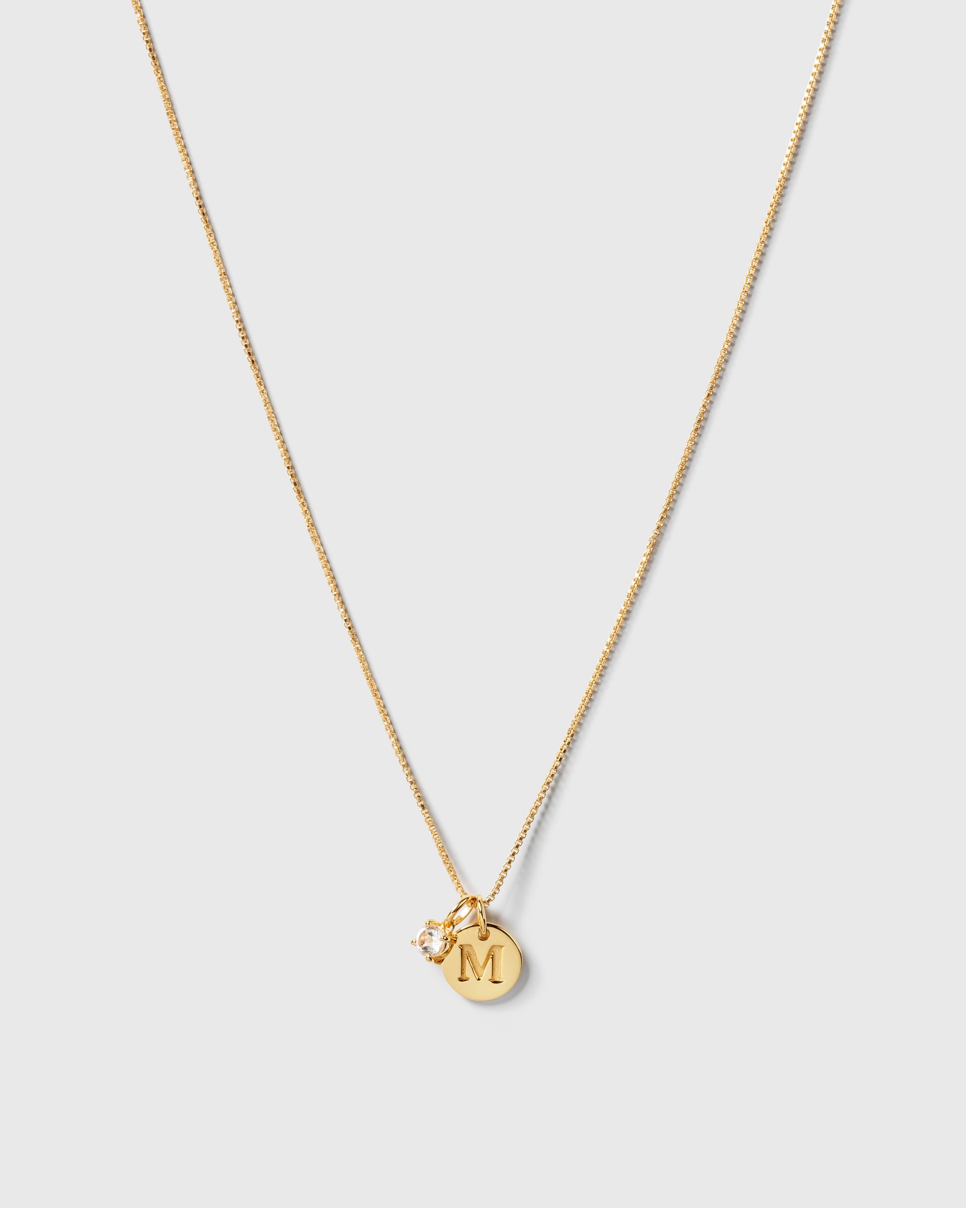 A product shot of a gold necklace featuring the Beloved Solo Nova Pendant with its white topaz stone, styled alongside a round gold pendant engraved with the letter 'M'.