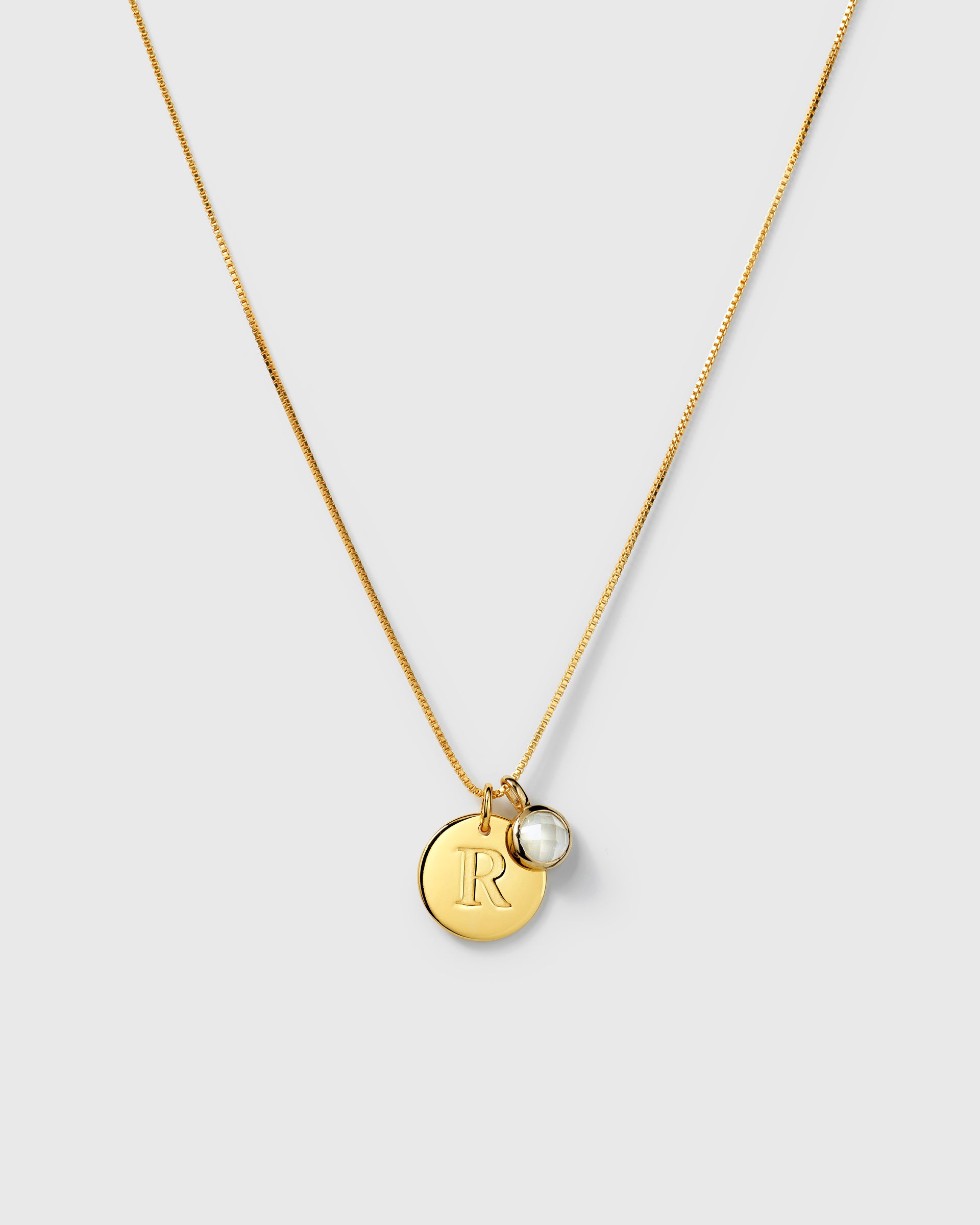 Gold box chain necklace with two pendants: a round letter charm and a round faceted moonstone. 