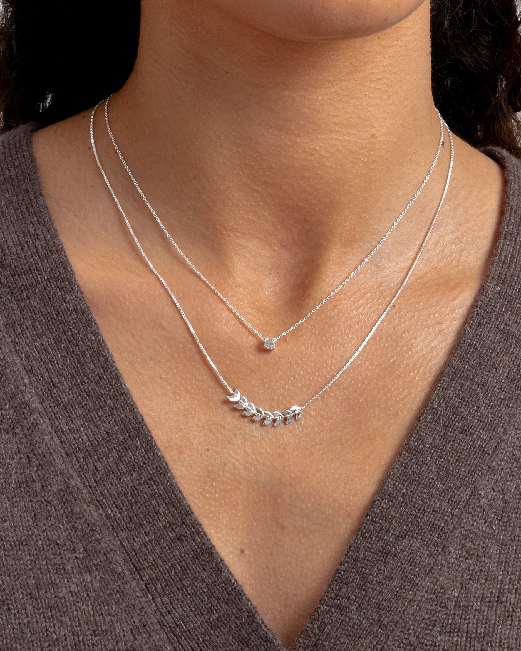 A silver necklace featuring delicate leaf-like details, layered with a shorter, fine silver chain adorned with a single clear stone.