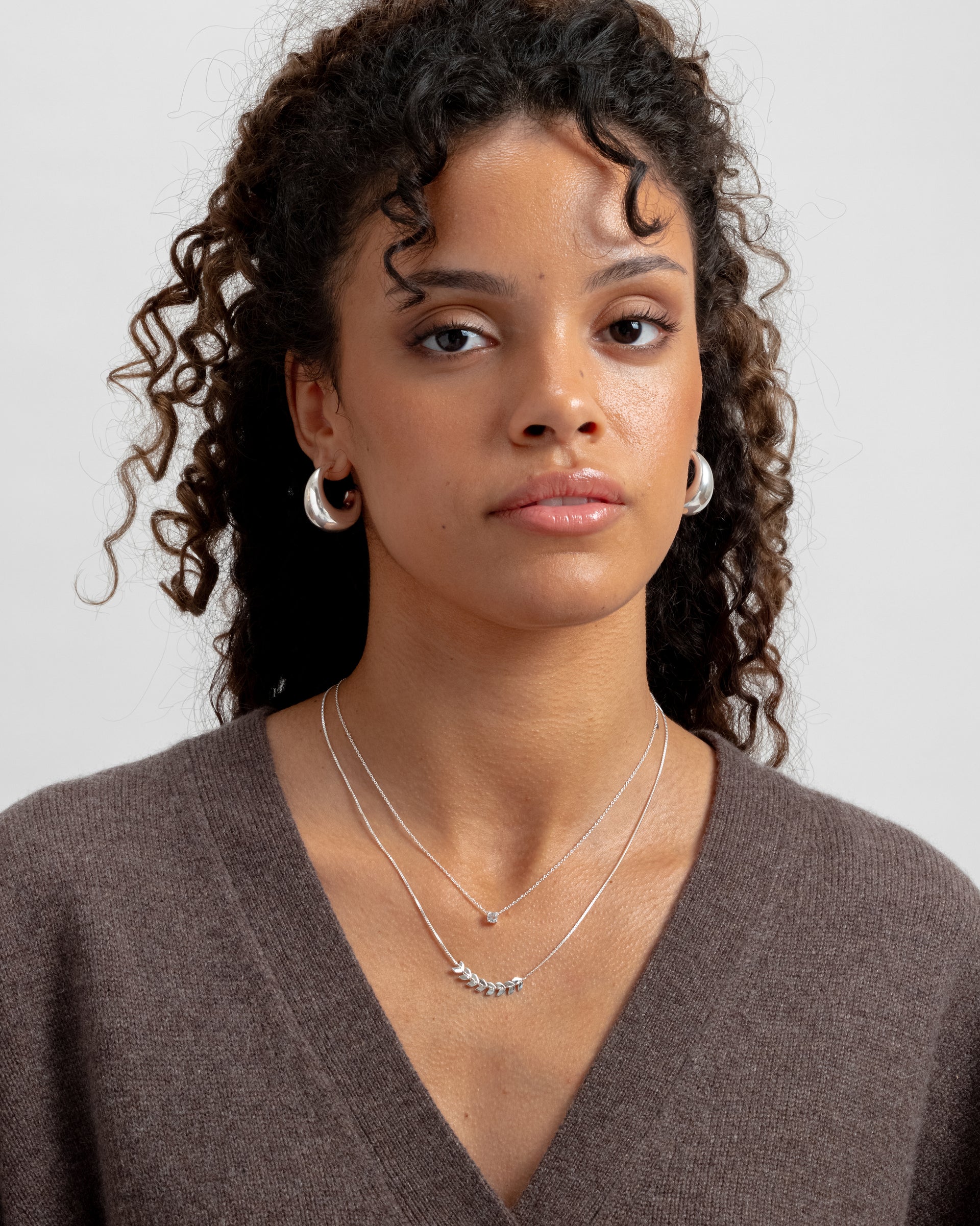 Layered silver necklace with delicate leaf-like details on the longer chain, paired with a shorter fine chain necklace, and chunky silver hoop earrings.