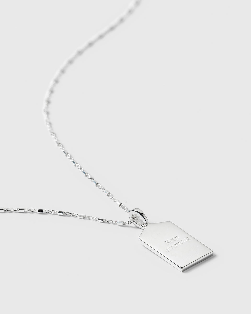 A close-up of the silver Lucky Coin Keep Dreaming Necklace, showing the back of the rectangular pendant engraved with the words 'Keep dreaming'.
