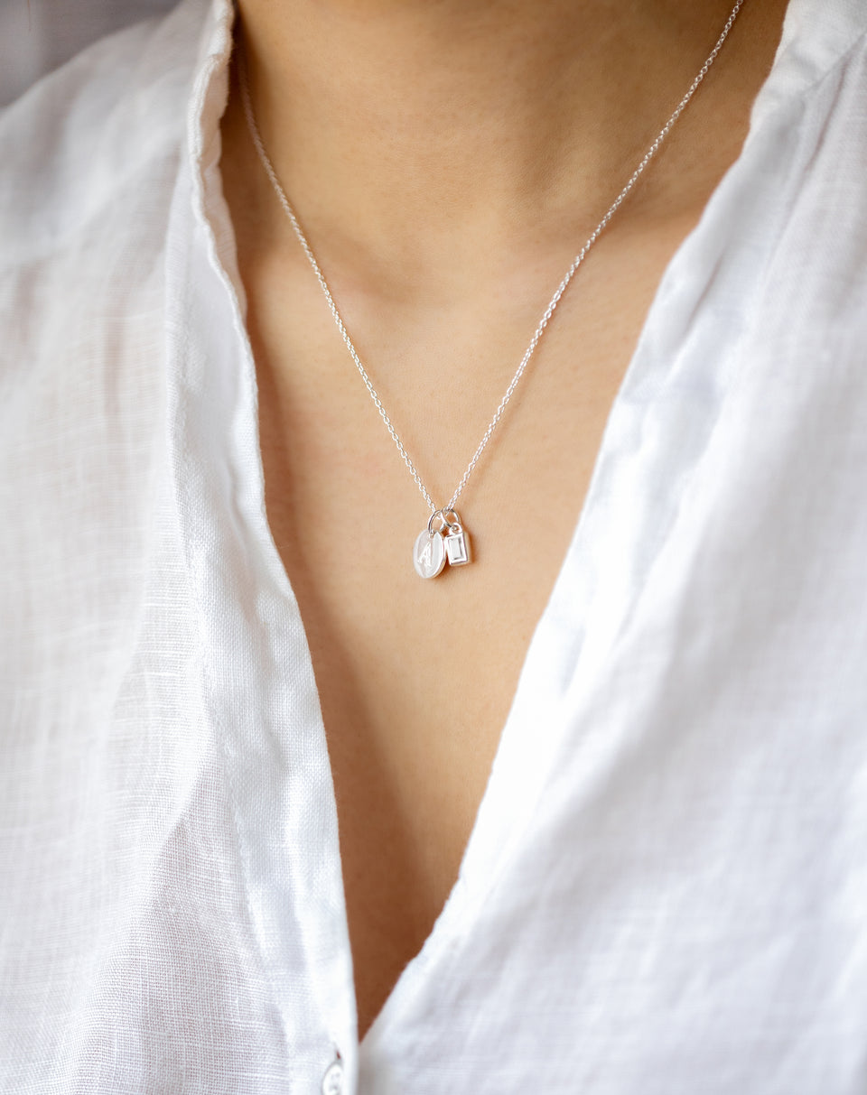 A close-up of a model wearing the silver Beloved Squared Pendant with a crystal, styled on a fine chain with a round letter charm and a small padlock charm.
