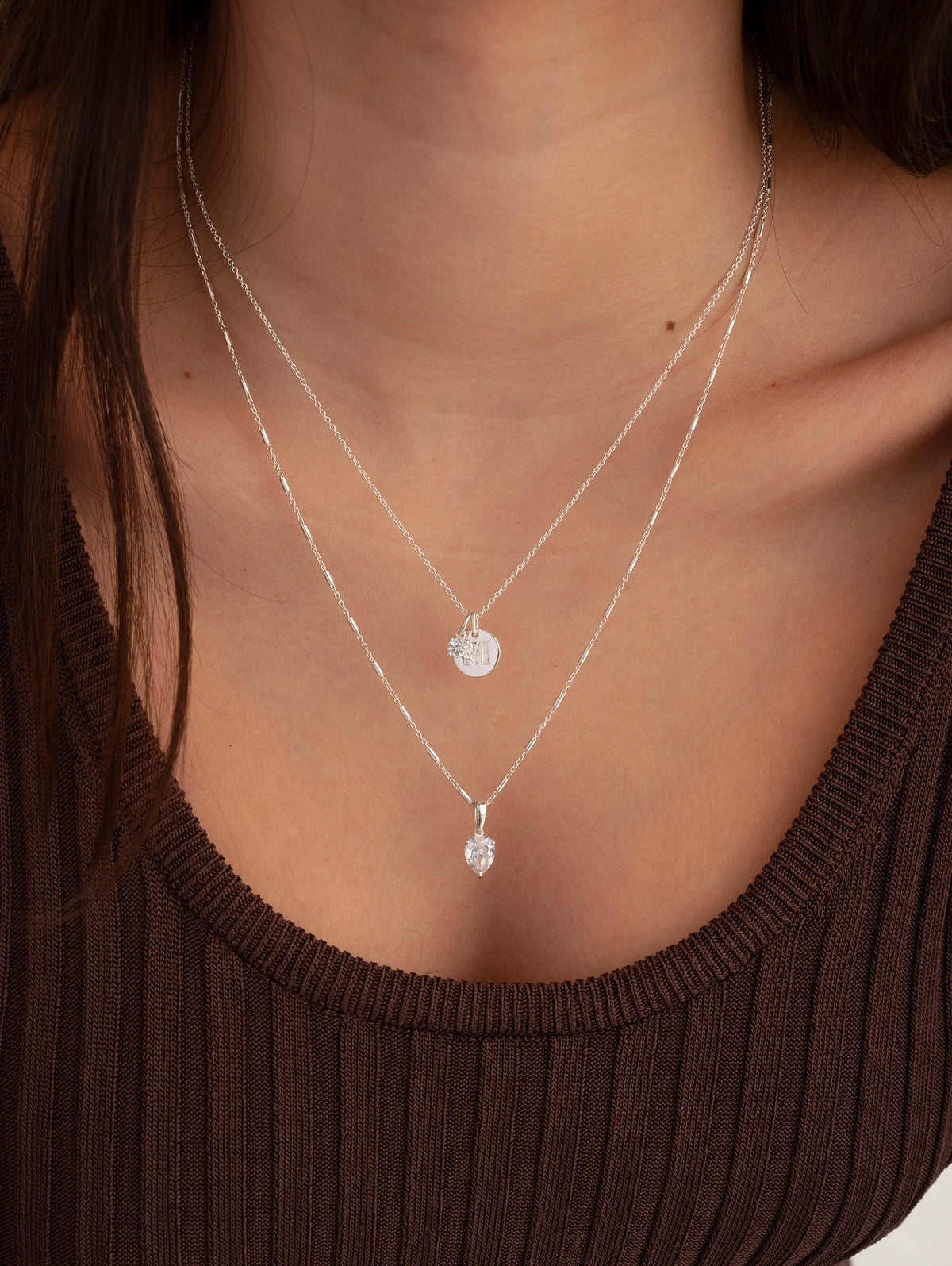 A model wears the Katie Necklace in silver, featuring a unique chain and a drop-shaped cubic zirconia pendant, layered with a second silver necklace that has a small, round engraved charm.