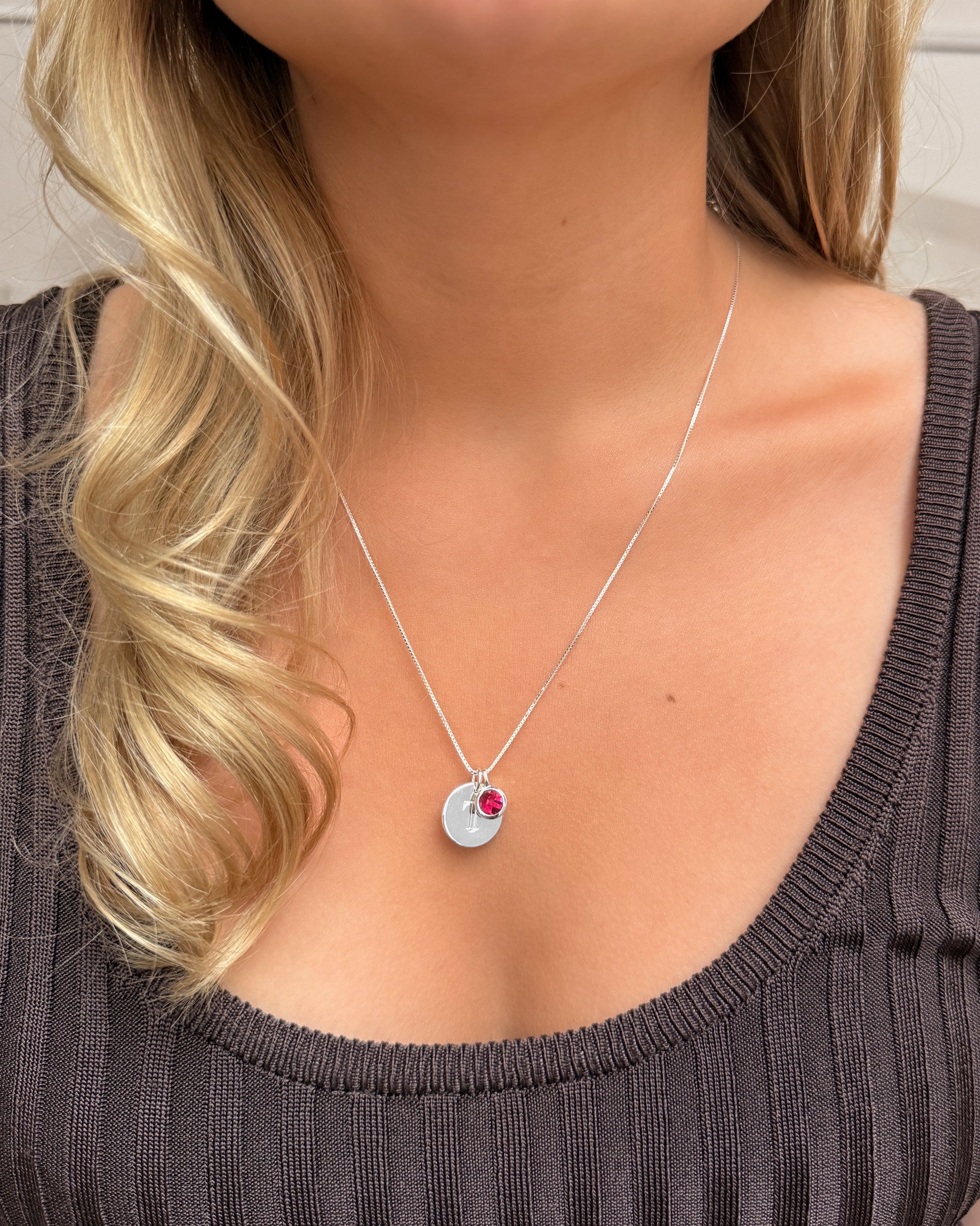 A silver necklace with a ruby pendant, July‚Äôs birthstone, layered with a delicate silver chain featuring a small letter pendant.
