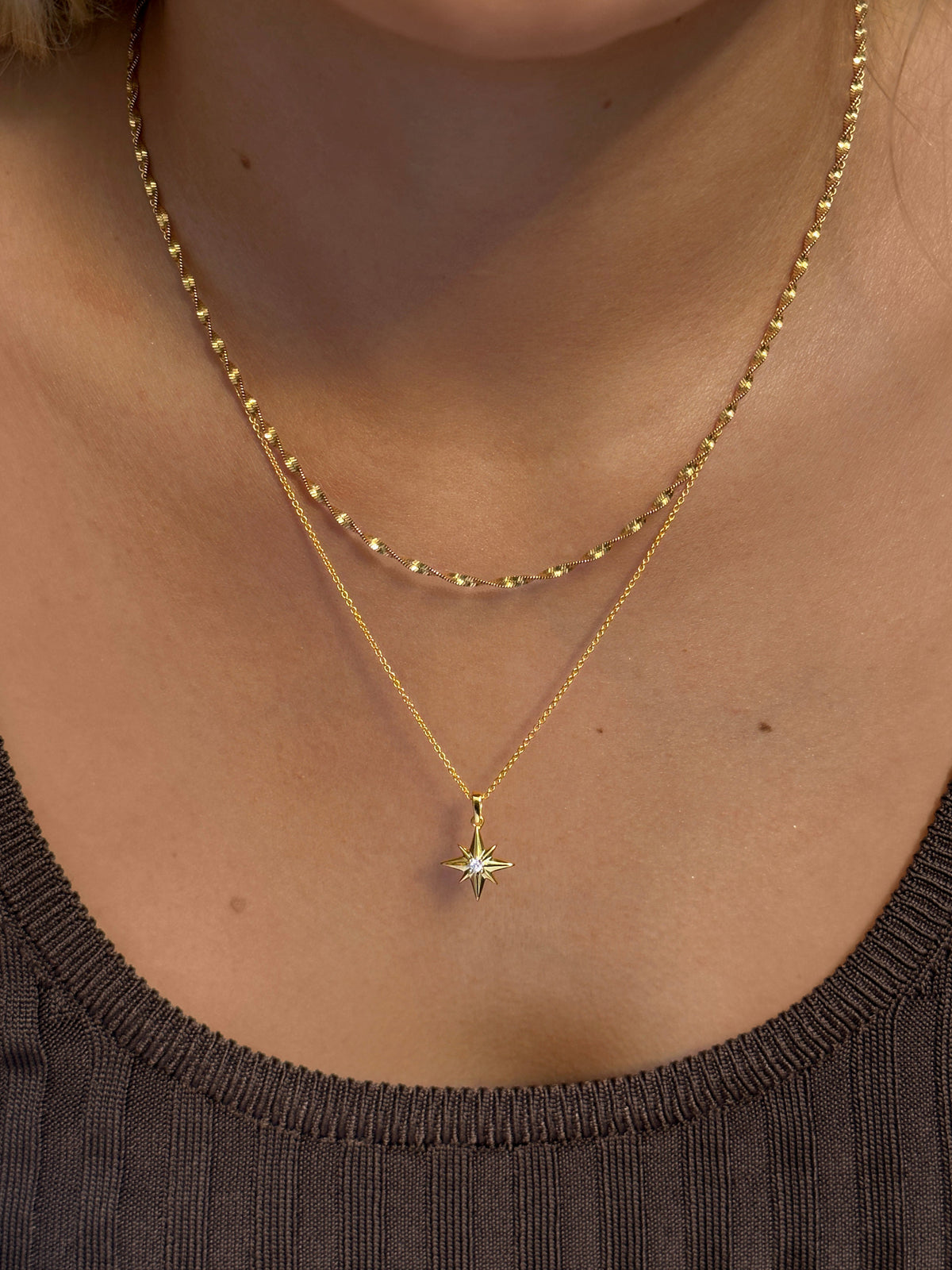 A close-up on a model's neck, showcasing a layered style with the gold North Star Big Necklace and a twisted gold chain. The North Star necklace features a delicate chain and a star-shaped pendant with a central crystal.