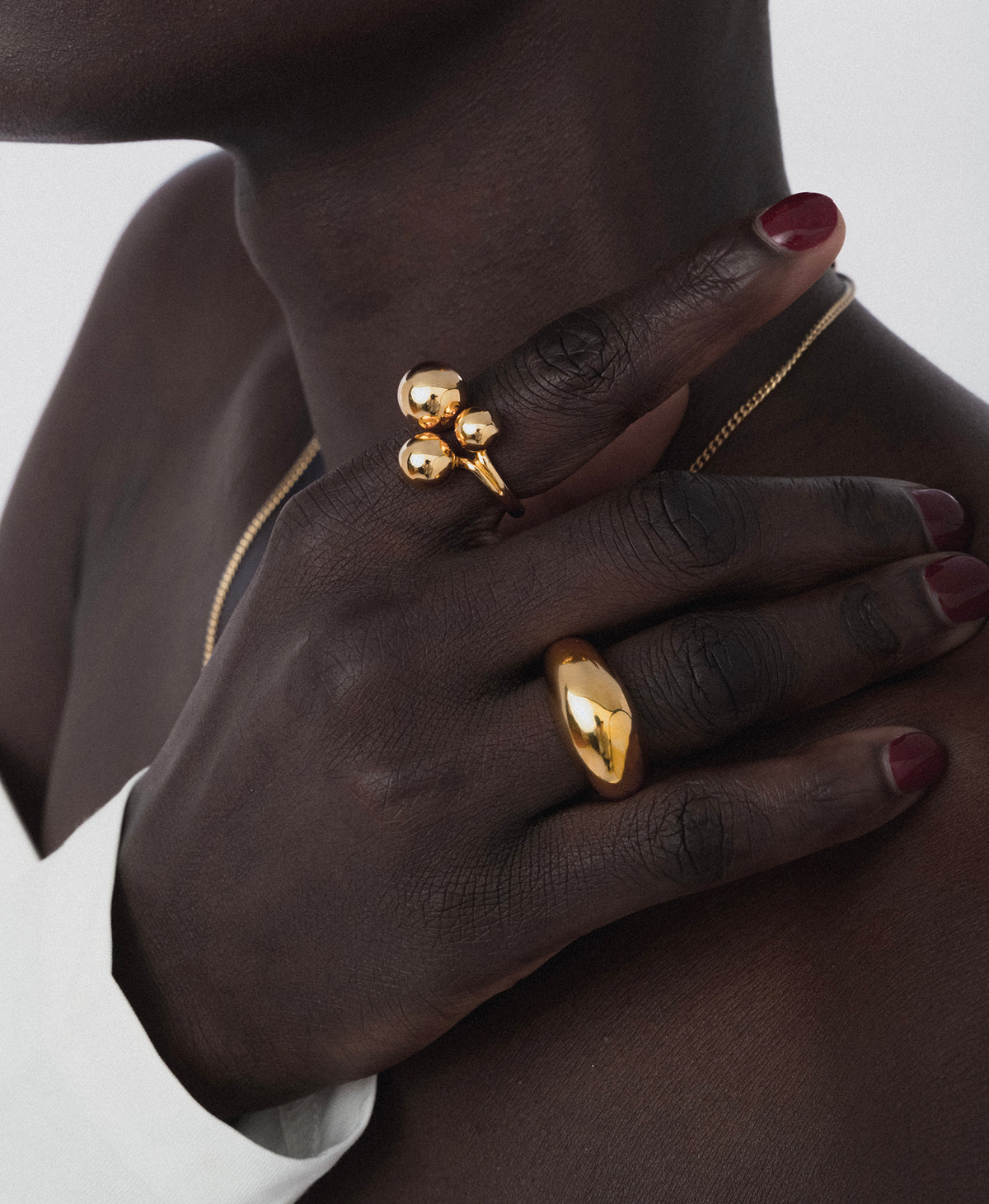 A close-up shot of a model's hand resting on her collarbone, showing off the Bolded Big Ring Gold on her ring finger. She is also wearing another gold ring with a three-sphere design on her index finger and a fine gold chain necklace.