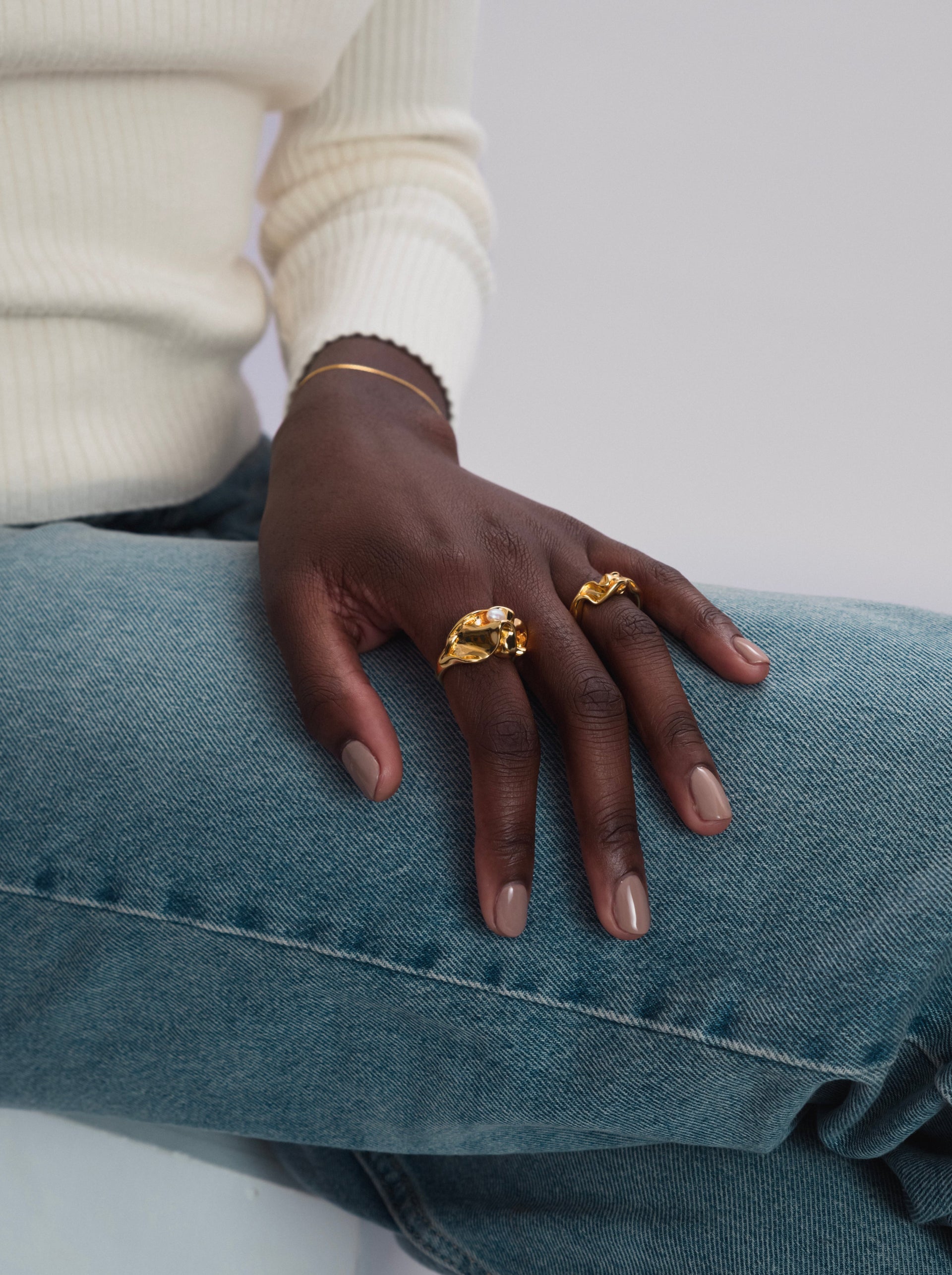 A close-up of a model wearing the Moulded Big Ring Gold, an irregularly shaped ring with a unique freshwater pearl, paired with a smaller wavy gold ring and a delicate gold bracelet.