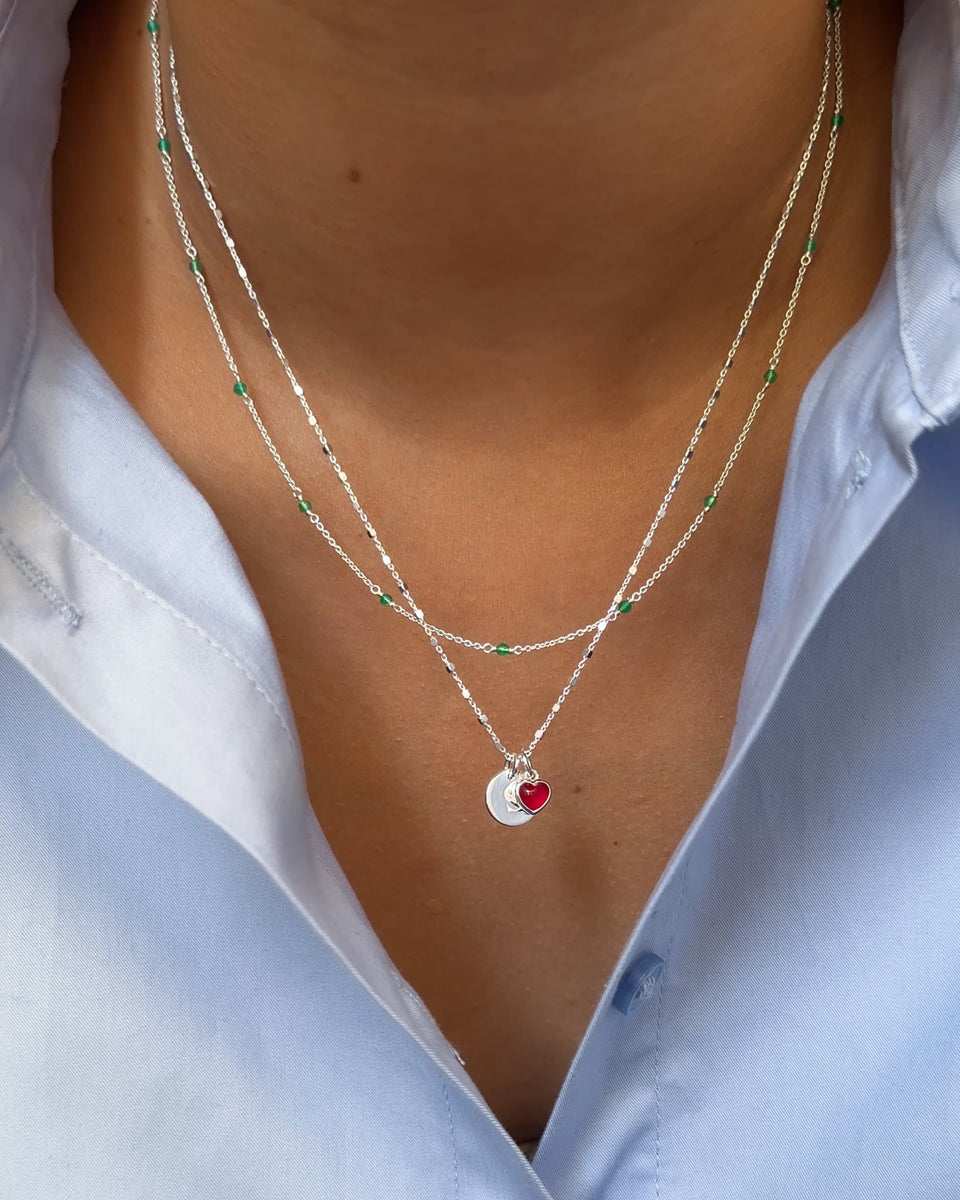 A close-up on a model's neck showing a layered necklace look featuring the silver Santa Monica Necklace with green onyx stones, the Sheer Station Necklace, and a shorter necklace with the Beloved Mini Letter and a pink Beloved Heart Pendant.