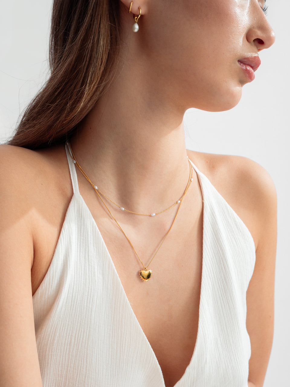 A model wearing a layered set of gold necklaces, the True Love Spike Heart Necklace, featuring a chain and a heart-shaped pendant with small spikes around its edge and the Treasure Multi Pearl Necklace. Complemented by the Treasure Pearl Hoops and Beloved Twisted Small Hoops.