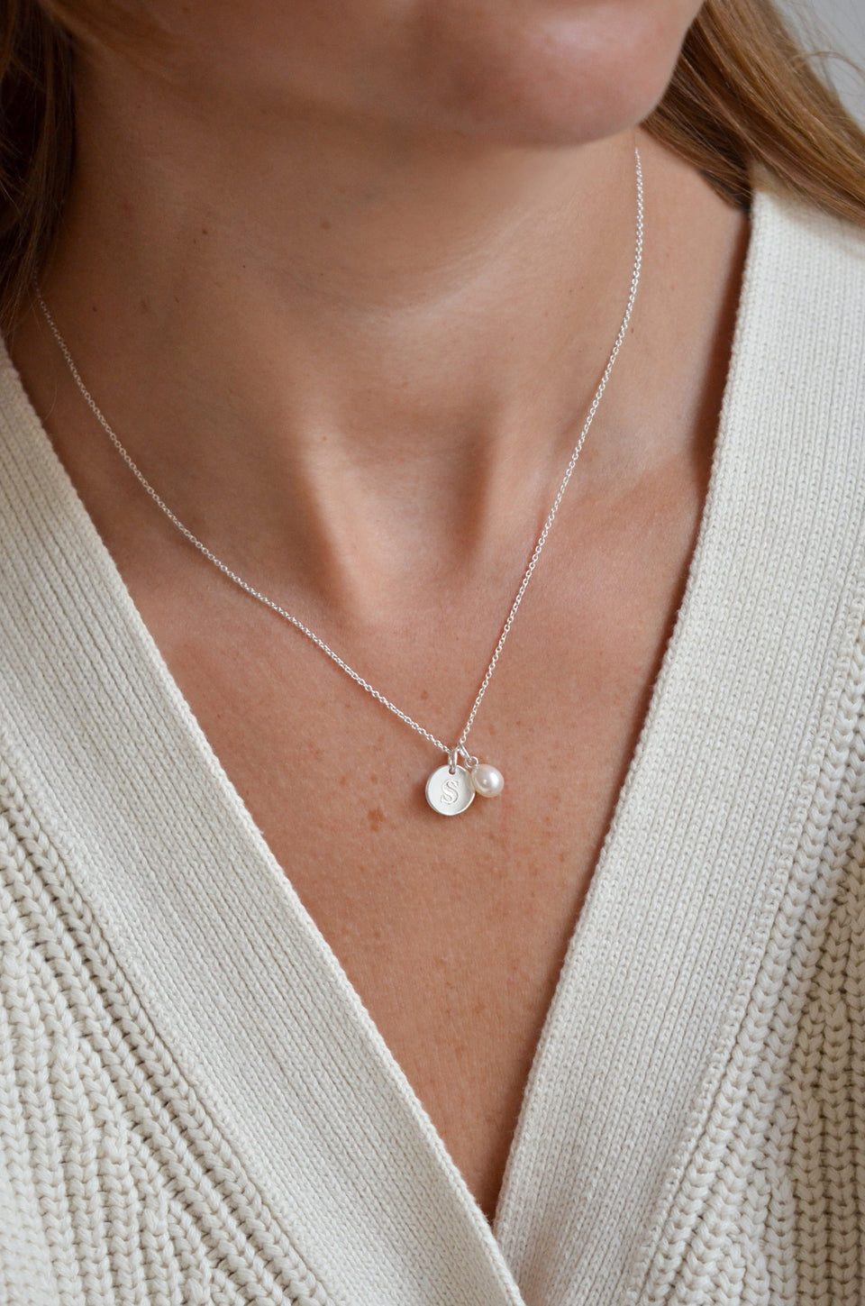 A close-up of a model wearing the Beloved Pearl Pendant in silver, styled on a delicate silver chain with an additional silver disc pendant engraved with the initial 'S'.